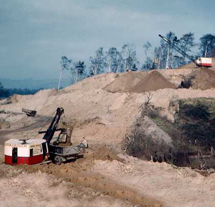 A view of the east portal looking north-west showing a 22RB face shovel loading a 5 cubic yard lorry. In the background, a 19RB dragline is forming the batters over the tunnel.